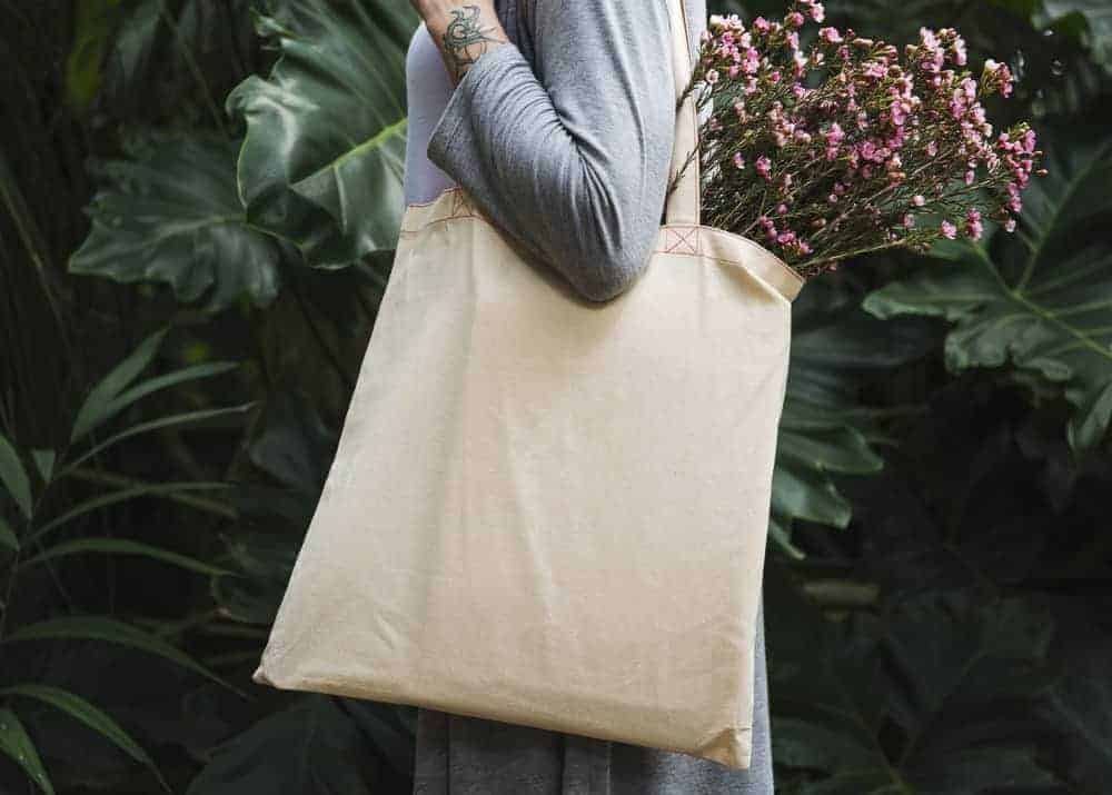 A person stands sideways facing the left in a gray long sleeved shirt holding a plain beige colored tote bag. We can only see their arm, chest, and the bottom part of their sweater. We can see their forearm and the wrist is uncovered and shows a piece of a black colored tattoo. The bag is holding small pink flowers. They are standing in front of big green leaves, which make up the entire background of the picture. 