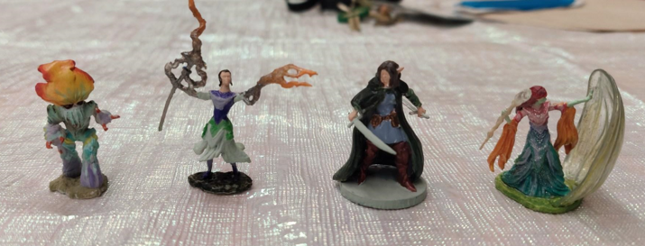a group of painted rpg miniature figures - a colorful mushroom character, a wizard doing magic, a rogue with a sword, and an elf wizard casting a spell.
