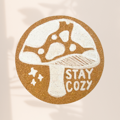 image of a tan cork coaster with a white mushroom and the words "stay cozy"